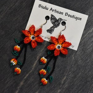 May include: Orange and yellow beaded flower dangle earrings with green leaves and black beads. The earrings are hanging from silver hooks. The earrings are on a brown wooden surface with a white card that says "Biulu Artisan Boutique".