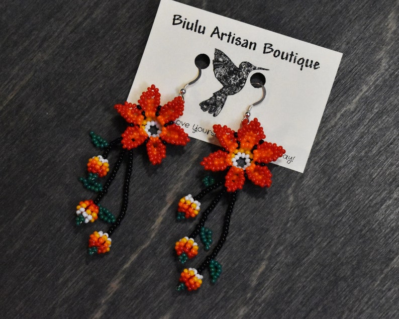 May include: Orange and yellow beaded flower dangle earrings with green leaves and black beads. The earrings are hanging from silver hooks. The earrings are on a brown wooden surface with a white card that says "Biulu Artisan Boutique".