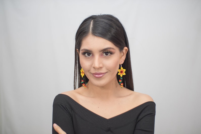 May include: A woman wearing yellow flower earrings with red and green accents. The earrings are dangling and have a beaded design. The woman is wearing a black off-the-shoulder top.