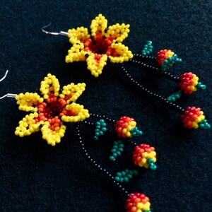 May include: A pair of beaded flower earrings. Each earring features a yellow and red flower with a black beaded stem. The stem has green and red beaded accents. The earrings are on a dark background.