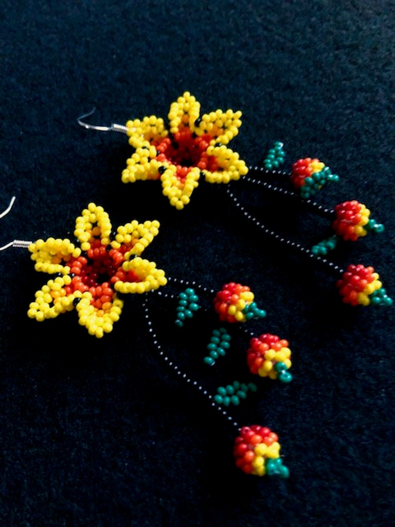 May include: A pair of beaded flower earrings. Each earring features a yellow and red flower with a black beaded stem. The stem has green and red beaded accents. The earrings are on a dark background.