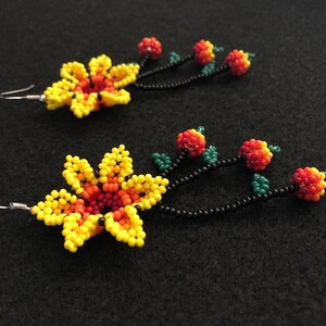 May include: A pair of beaded earrings featuring a yellow and orange flower design with a black stem and red and orange beaded berries. The earrings have silver-colored ear wires and are set against a black background.