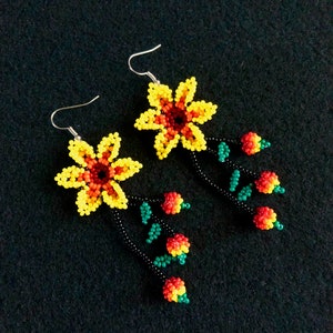May include: A pair of beaded earrings featuring yellow and orange flower designs with dangling red and green beaded accents. The earrings have silver-colored hooks and are set against a black background. The floral design is intricate and colorful.