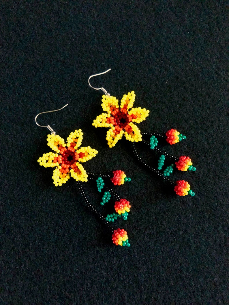 May include: A pair of beaded earrings featuring yellow and orange flower designs with dangling red and green beaded accents. The earrings have silver-colored hooks and are set against a black background. The floral design is intricate and colorful.