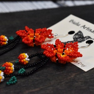 May include: A pair of beaded earrings with a flower design. The earrings are made with red, orange, white, green, and black beads. The earrings are hanging from silver hooks.