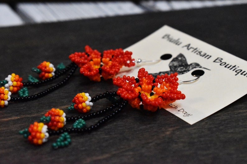 May include: A pair of beaded earrings with a flower design. The earrings are made with red, orange, white, green, and black beads. The earrings are hanging from silver hooks.