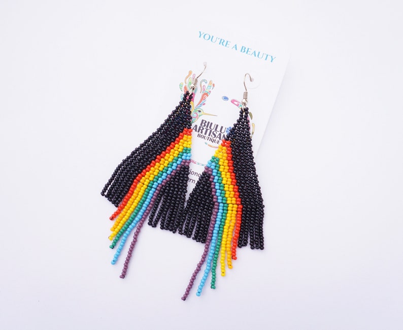 May include: A pair of beaded earrings with a rainbow design. The earrings are black with a rainbow pattern of red, orange, yellow, green, blue, and purple beads. The earrings are hanging from a white card that says "YOU'RE A BEAUTY".
