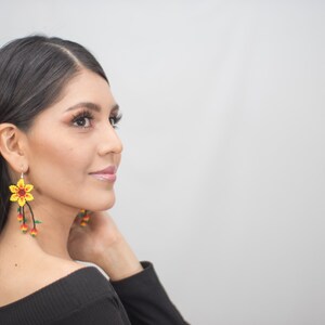 May include: Yellow beaded flower earrings with red and green accents. The earrings have a floral design with dangling beaded strands. The earrings are worn by a person with dark hair and a black top.