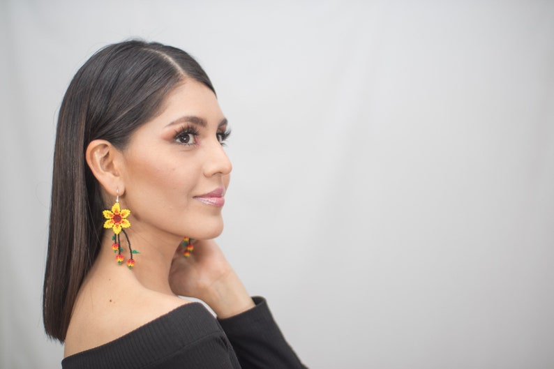 May include: Yellow beaded flower earrings with red and green accents. The earrings have a floral design with dangling beaded strands. The earrings are worn by a person with dark hair and a black top.