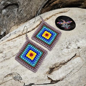 May include: A pair of square-shaped earrings with a colourful beaded design. The earrings are made with small, colourful beads in a geometric pattern. The earrings are a light purple colour and have silver hooks.