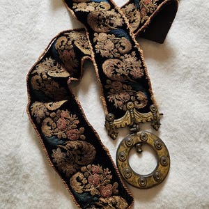 May include: A decorative black belt featuring a floral design in gold, pink, and blue. It has a metal clasp and a circular buckle. The edges are trimmed with a thin, brown border. The belt is likely vintage or antique, with a detailed pattern.