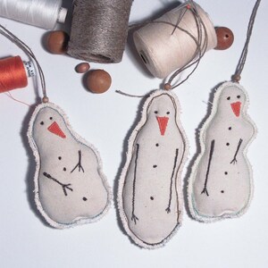 Puede incluir: Tres adornos de muñecos de nieve hechos a mano con picos naranjas y ojos y brazos de botón negros. Los muñecos de nieve están hechos de tela blanquecina con un borde deshilachado y cuelgan de una cuerda. Bobinas de hilo y cuentas de madera en el fondo.