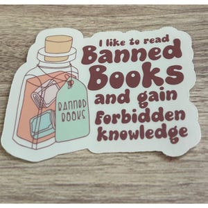 May include: A sticker featuring a jar with a cork, containing a book and a tag that reads "Banned Books." The sticker also has the text "I like to read Banned Books and gain forbidden knowledge" in maroon.