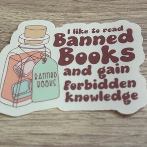 May include: A sticker with a jar illustration containing books and a tag that says "Banned Books." The sticker has text that reads "I like to read Banned Books and gain forbidden knowledge."