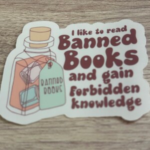 May include: A sticker featuring a jar with books inside and a tag that reads "Banned Books." The text on the sticker says, "I like to read Banned Books and gain forbidden knowledge." The sticker has a white border.