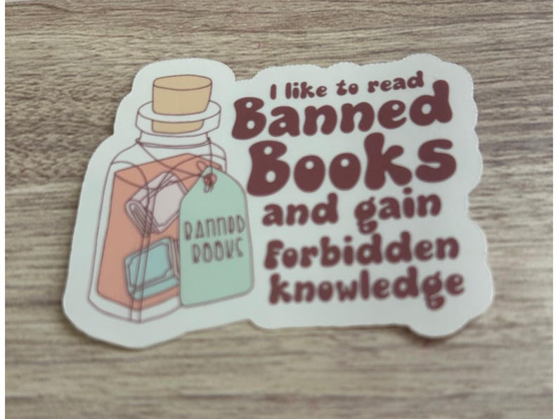May include: A sticker featuring a jar with books inside and a tag that reads "Banned Books." The text on the sticker says, "I like to read Banned Books and gain forbidden knowledge." The sticker has a white border.
