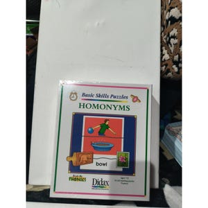 May include: A boxed educational puzzle set titled "Basic Skills Puzzles HOMONYMS." The box features a colorful illustration of a person throwing a ball into a bowl, with the word "bowl" below. The puzzle is for ages 7-9.