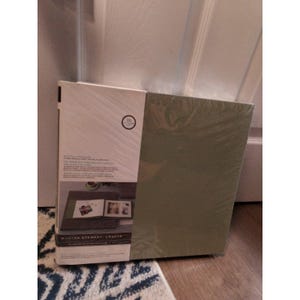 May include: A Martha Stewart Crafts photo album in a sage green color, partially wrapped in clear plastic. The album's packaging features a circular design with the number 10 and product information. The album is displayed on a wooden surface.