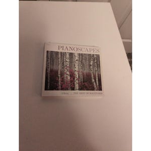 May include: A CD case with the title "PIANOSCAPES" in white letters. The cover features a photograph of a forest with tall, white birch trees and pink foliage. The text "THE BEST OF SOLITUDES" is at the bottom.