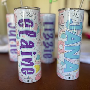 May include: Three white stainless steel tumblers with colorful cartoon designs and names "Jana", "Olivia" and "Lizzie" printed on them. Each tumbler has a clear plastic lid and a colorful straw.