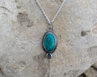 May include: A silver chain necklace with a pendant featuring a teal oval gemstone set in a silver bezel. The pendant has a small silver bead hanging from the bottom.
