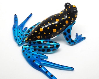 May include: A frog figurine featuring a glossy black back with bright yellow spots. The limbs are a vibrant blue with black spots. The frog is depicted in a crouching position with its legs extended outward, showcasing a smooth, reflective surface.