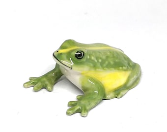 May include: A small ceramic frog figurine with a green and yellow color scheme. The frog is sitting on a white surface.