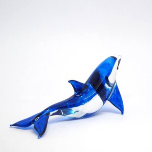 May include: A blue glass sculpture of an orca whale with a white underbelly. The sculpture is posed in a swimming position with its tail raised.