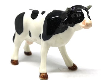 May include: A ceramic cow figurine with a black and white spotted pattern. The cow has a black head and back, with white legs and body. The hooves are brown, and the figurine is standing on a white surface.
