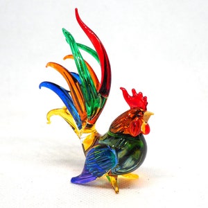 Puede incluir: Una figura de gallo de cristal con un dise&ntilde;o multicolor vibrante. El cuerpo est&aacute; hecho de vidrio transl&uacute;cido verde y azul, mientras que la cresta y las barbillas son de color rojo brillante. Las plumas de la cola son alargadas, curvas y de colores rojo, verde, azul, naranja y amarillo.