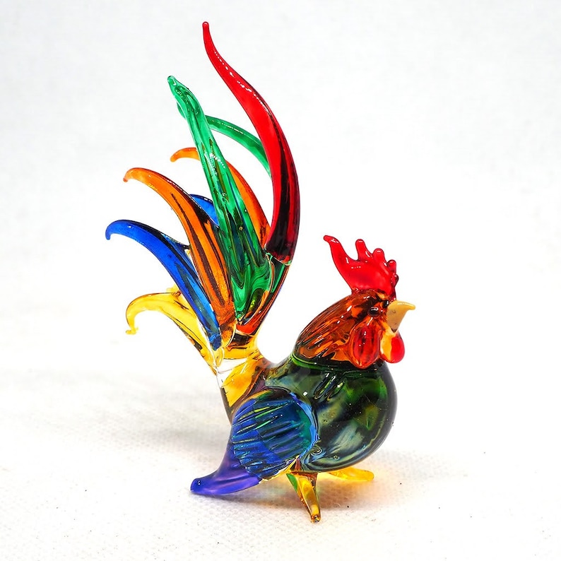 Puede incluir: Una figura de gallo de cristal con un dise&ntilde;o multicolor vibrante. El cuerpo est&aacute; hecho de vidrio transl&uacute;cido verde y azul, mientras que la cresta y las barbillas son de color rojo brillante. Las plumas de la cola son alargadas, curvas y de colores rojo, verde, azul, naranja y amarillo.