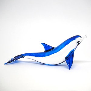 May include: A blue and white glass dolphin figurine. The dolphin is swimming with its head turned to the side and its tail fin extended.