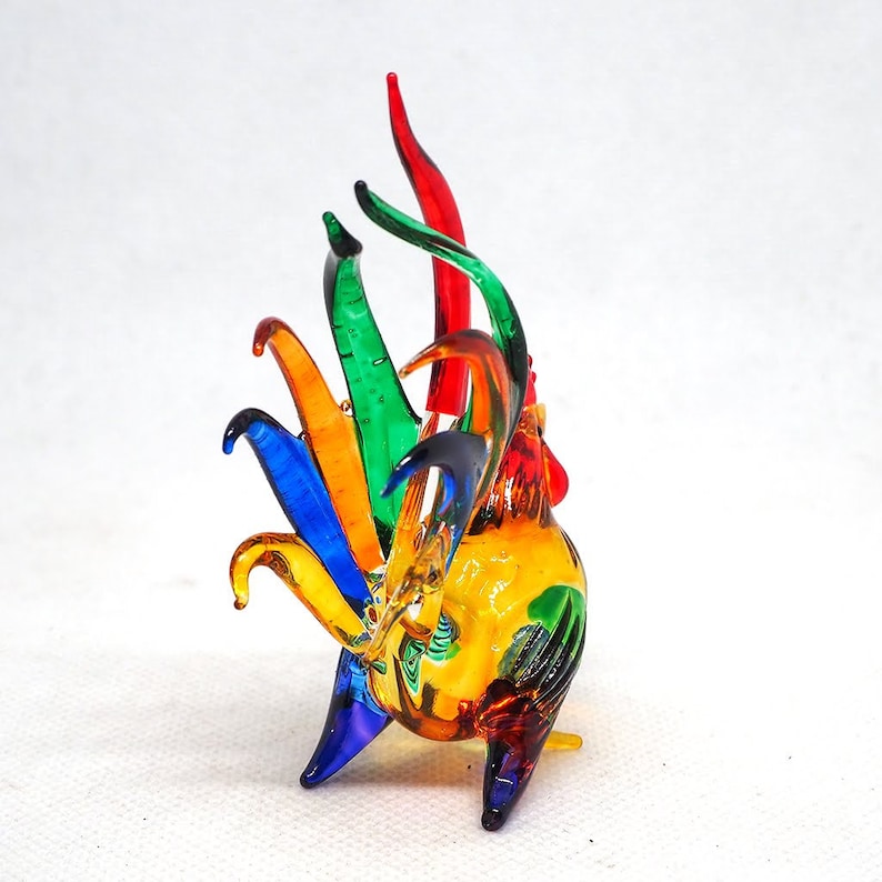 Puede incluir: Una figura de gallo de vidrio multicolor que presenta una cola vibrante con plumas alargadas y curvas en tonos azul, naranja, verde y rojo. El cuerpo est&aacute; elaborado con vidrio amarillo transl&uacute;cido con acentos en rojo y verde, con acabado liso.