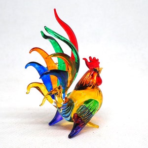 Puede incluir: Una figura de gallo de vidrio hecha a mano con un dise&ntilde;o multicolor vibrante. El cuerpo es principalmente amarillo con alas verdes, mientras que las plumas de la cola muestran un espectro de rojo, verde, naranja, azul y amarillo. Cresta roja.