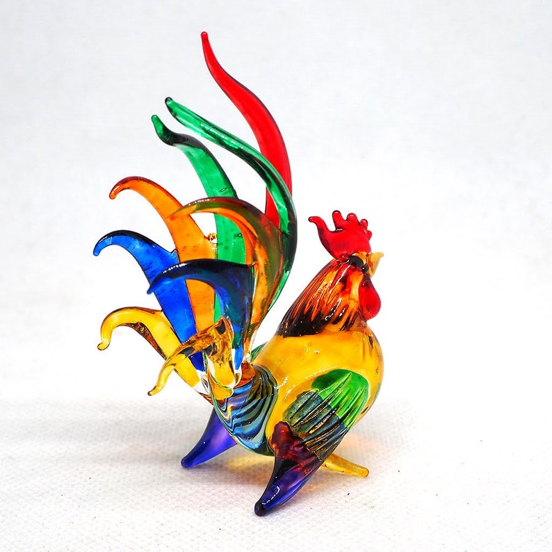 Puede incluir: Una figura de gallo de vidrio hecha a mano con un dise&ntilde;o multicolor vibrante. El cuerpo es principalmente amarillo con alas verdes, mientras que las plumas de la cola muestran un espectro de rojo, verde, naranja, azul y amarillo. Cresta roja.