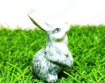 May include: A small ceramic rabbit figurine with a glossy finish, featuring a white base coat with grey mottled markings across its body, upright ears, and a small orange-toned nose, depicted in a seated position with its front paws tucked in.