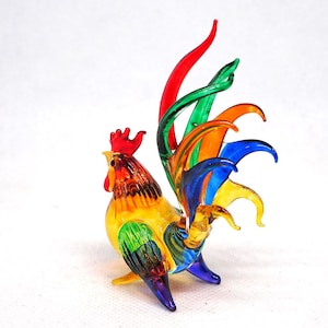Puede incluir: Una figura de gallo de cristal con un dise&ntilde;o multicolor vibrante. El cuerpo muestra tonos amarillos, naranjas y verdes, mientras que las plumas de la cola est&aacute;n elaboradas con hebras alargadas y curvas de cristal rojo, verde, naranja, azul y amarillo.
