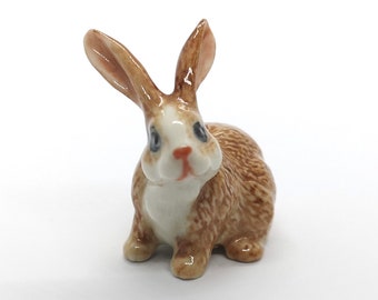 May include: A small ceramic figurine of a brown and white rabbit with blue eyes. The rabbit is sitting with its ears up and its head tilted slightly to the side.