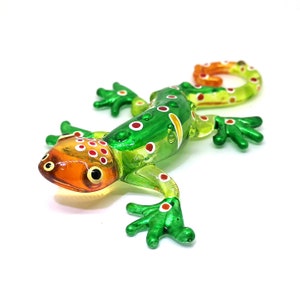 May include: A green glass gecko figurine with orange and white accents. The gecko has a curled tail and is decorated with small white dots.
