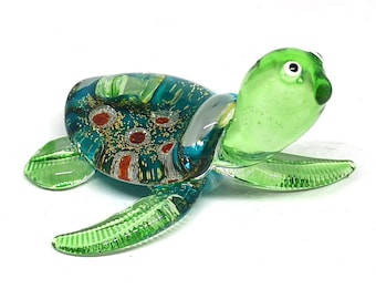 Puede incluir: Una figura de tortuga marina de vidrio con cabeza y aletas de color verde claro translúcido. El caparazón es de un azul cerceta profundo con motas de purpurina dorada incrustadas y patrones circulares en tonos naranja y blanco.