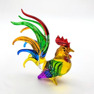 May include: A multi-colored glass rooster figurine featuring a vibrant red comb, a yellow body with green accents, and a long, flowing tail made of translucent red, green, yellow, and blue glass strands that curve elegantly upward.