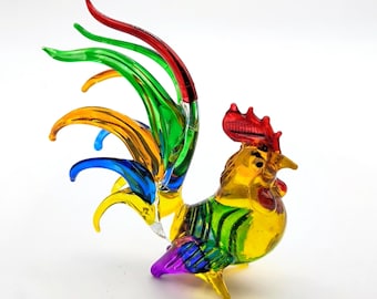 May include: A multi-colored glass rooster figurine featuring a vibrant red comb, a yellow body with green accents, and a long, flowing tail made of translucent red, green, yellow, and blue glass strands that curve elegantly upward.