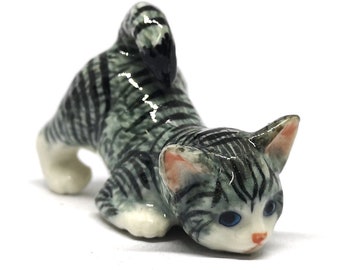 May include: A ceramic figurine of a gray and white tabby cat in a playful pose. The cat has blue eyes, pink ears, and a pink nose. The figurine is detailed with black stripes and white paws.