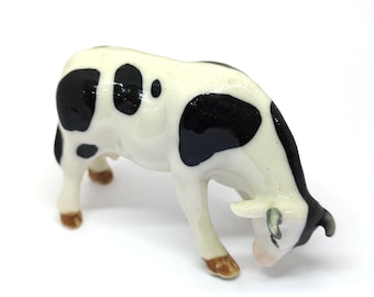 May include: A white and black ceramic cow figurine with black spots. The cow is in a grazing position with its head down.