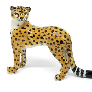 May include: A ceramic cheetah figurine with a tan body, black spots, and a white underbelly. The cheetah has a black and white striped tail and is posed in a standing position. The figurine has yellow eyes and is set against a white background.