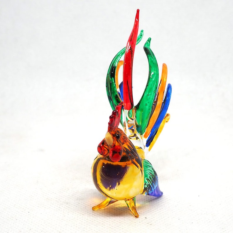 Puede incluir: Una figura de gallo de vidrio hecha a mano con un dise&ntilde;o vibrante y multicolor. El cuerpo es principalmente de color &aacute;mbar y amarillo, con una cresta y barbillas de color rojo brillante. La cola consta de plumas de vidrio alargadas y curvas en verde, rojo, naranja y azul.