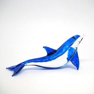 May include: A blue glass dolphin figurine with a white belly. The dolphin is swimming with its head tilted up and its tail fin extended.