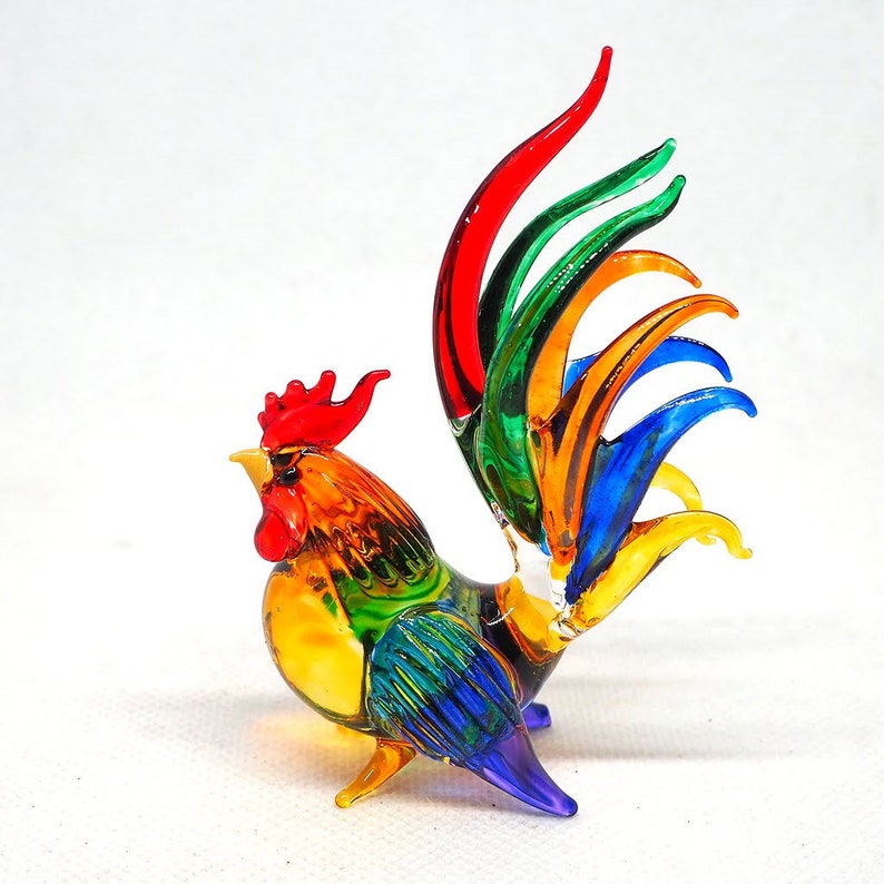 Puede incluir: Una figura de gallo de cristal con un dise&ntilde;o multicolor vibrante. El cuerpo muestra tonos amarillos, naranjas y verdes, mientras que las plumas de la cola est&aacute;n elaboradas en segmentos alargados y curvos de vidrio rojo, verde, naranja y azul.