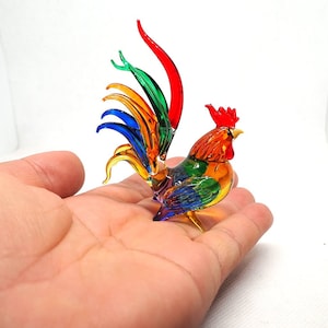 Puede incluir: Una peque&ntilde;a figura de gallo de vidrio multicolor que presenta una cresta roja vibrante, un pico amarillo y una cola compuesta por plumas de vidrio alargadas y curvas en tonos rojo, verde, naranja y azul brillantes, con un acabado transl&uacute;cido.