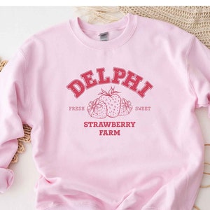 Op de afbeelding: Roze sweatshirt met een rood grafisch ontwerp met de tekst "DELPHI FRESH SWEET STRAWBERRY FARM" en een tekening van drie aardbeien.